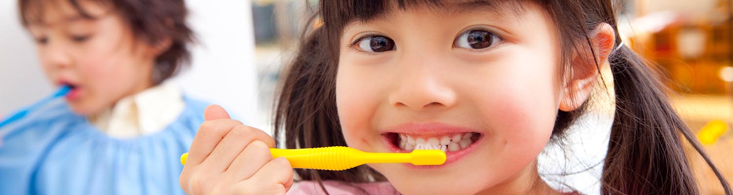 smiling asian preschooler smiling with yellow toothbrush on her teeth, blurred boy in background also brushing his teeth