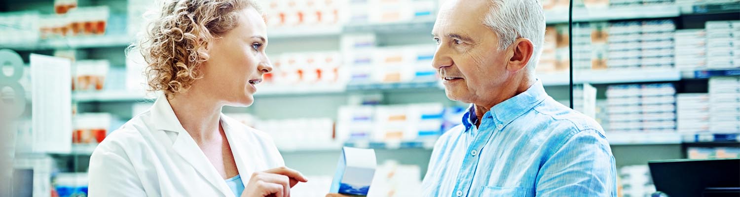 female pharmacist pointing to medication and talking with a senior male patient , pharmacy wall with medicine seen in background