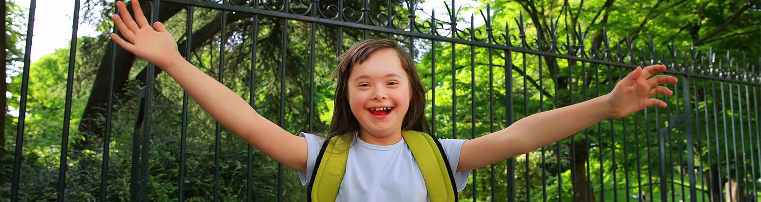 young girl with down syndrome and long brown hair, wearing backpack and smiling in park with beautiful green trees