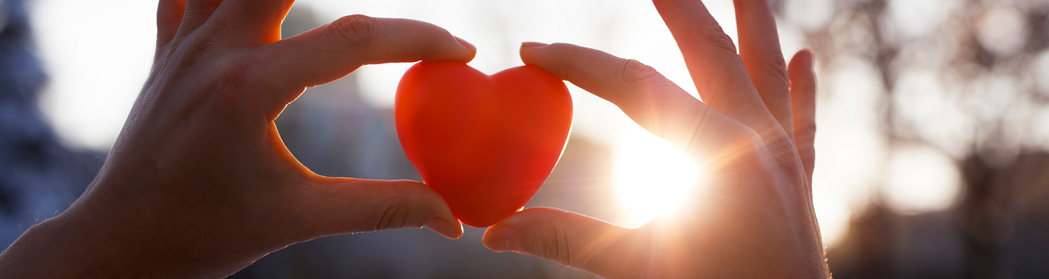 Closeup of woman's hands holding a red plastic heart, with sun light surronding