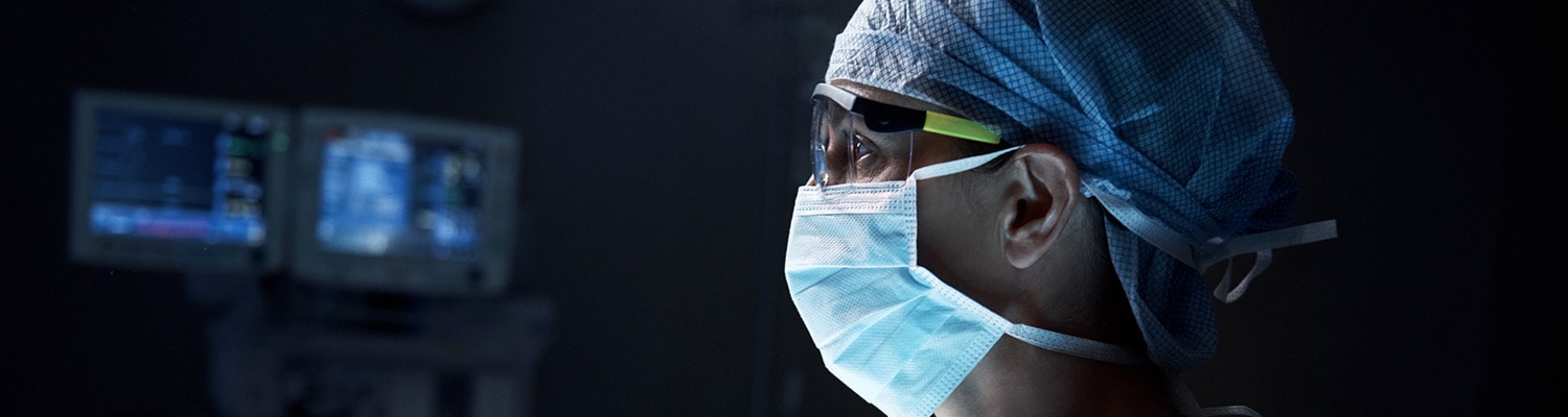 Surgeon in dark operating room with mask and protective glasses on looking at monitor.