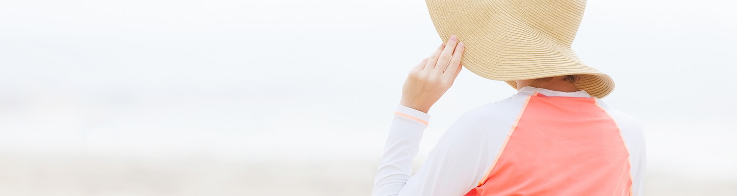 Back of woman at beach demonstrating sun protection with a floppy hat and a long sleeve shirt.