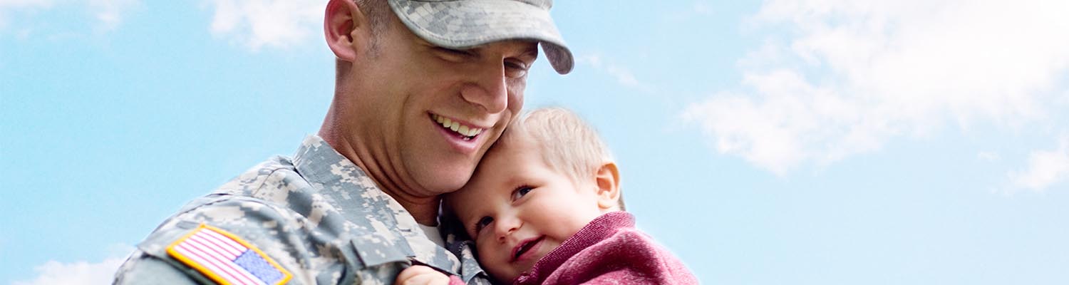 man in fatigues with american flag on shoulder  and a camouflage hat, smiling and holding toddler boy who is smiling and resting his head on his shoulders