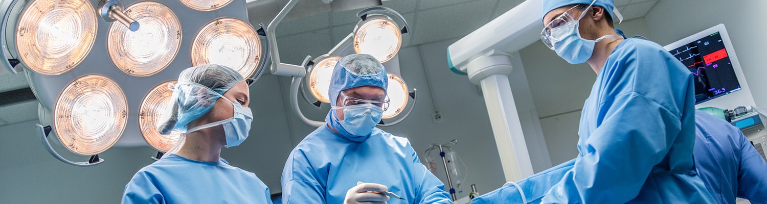 Surgeons in blue scrubs and masks in operating room with bright overhead lights.